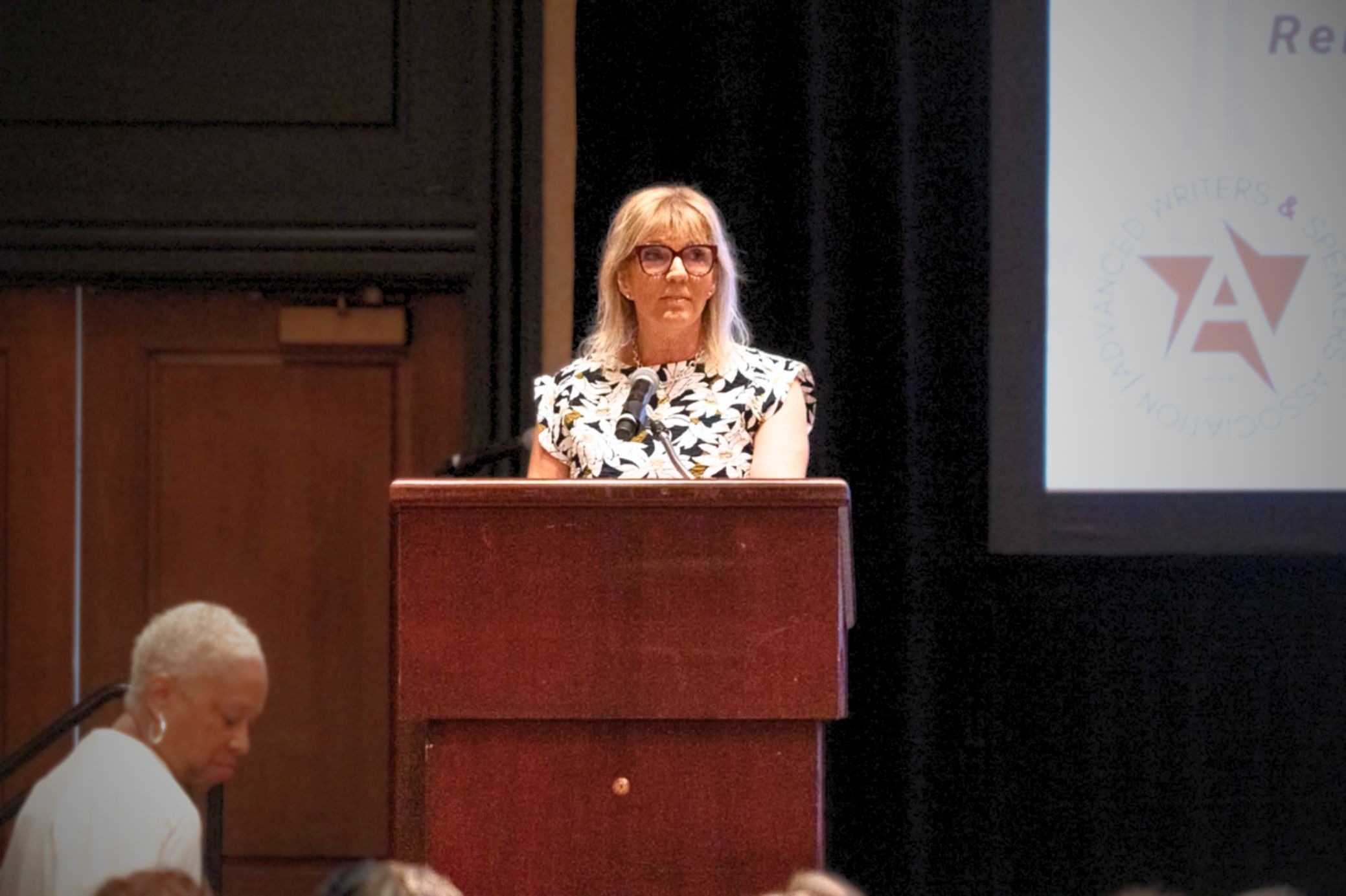 Blonde woman wearing glasses at a wooden podium delivering a talk, microphone in front, audience in view